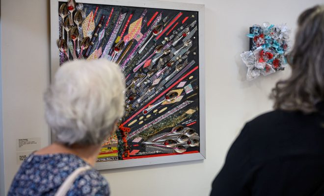 Two gallery visitors viewing framed mixed-media artwork on display.