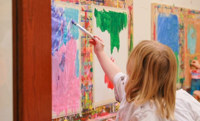 A young child holding a paintbrush and applying bright paint to a colorful art canvas.