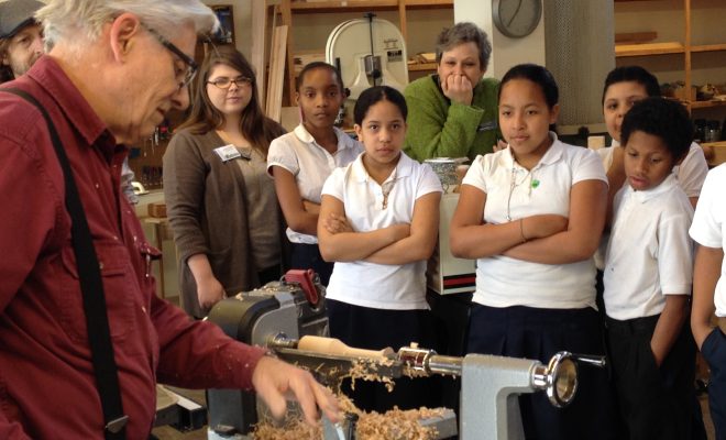 An instructor demonstrates woodturning on a lathe to a group of students in a workshop.