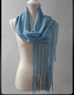 A light blue woven scarf with fringe styled on a clothing mannequin.