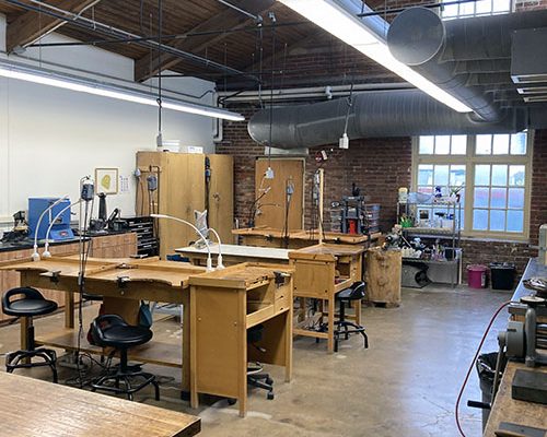 Spacious metals and jewelry studio with multiple wooden workbenches, tools, and large windows.