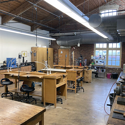 Spacious metals and jewelry studio with multiple wooden workbenches, tools, and large windows.