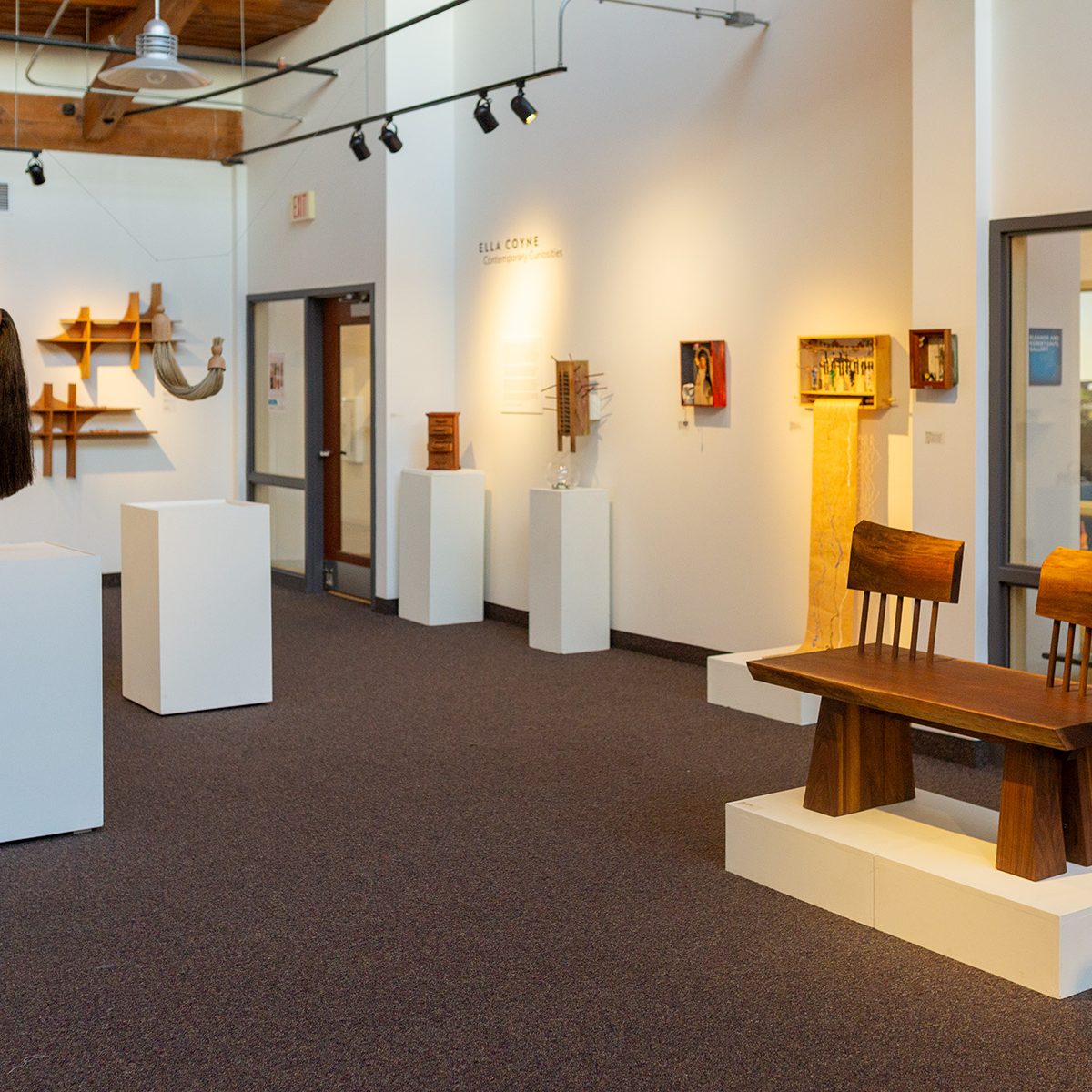 An art gallery exhibition displaying wooden furniture and wall sculptures on white pedestals.