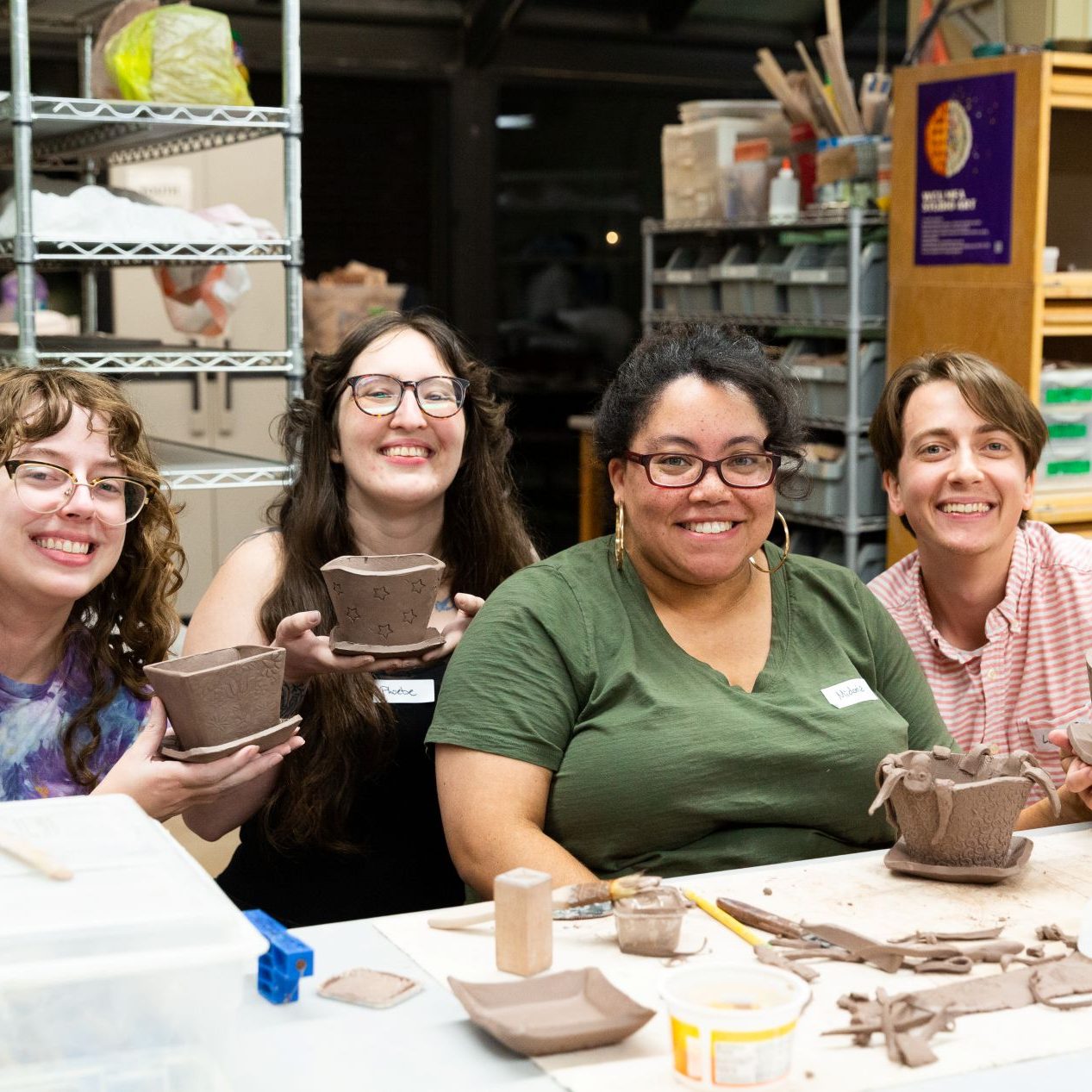 Four smiling women displaying their clay pots at a pottery studio table.