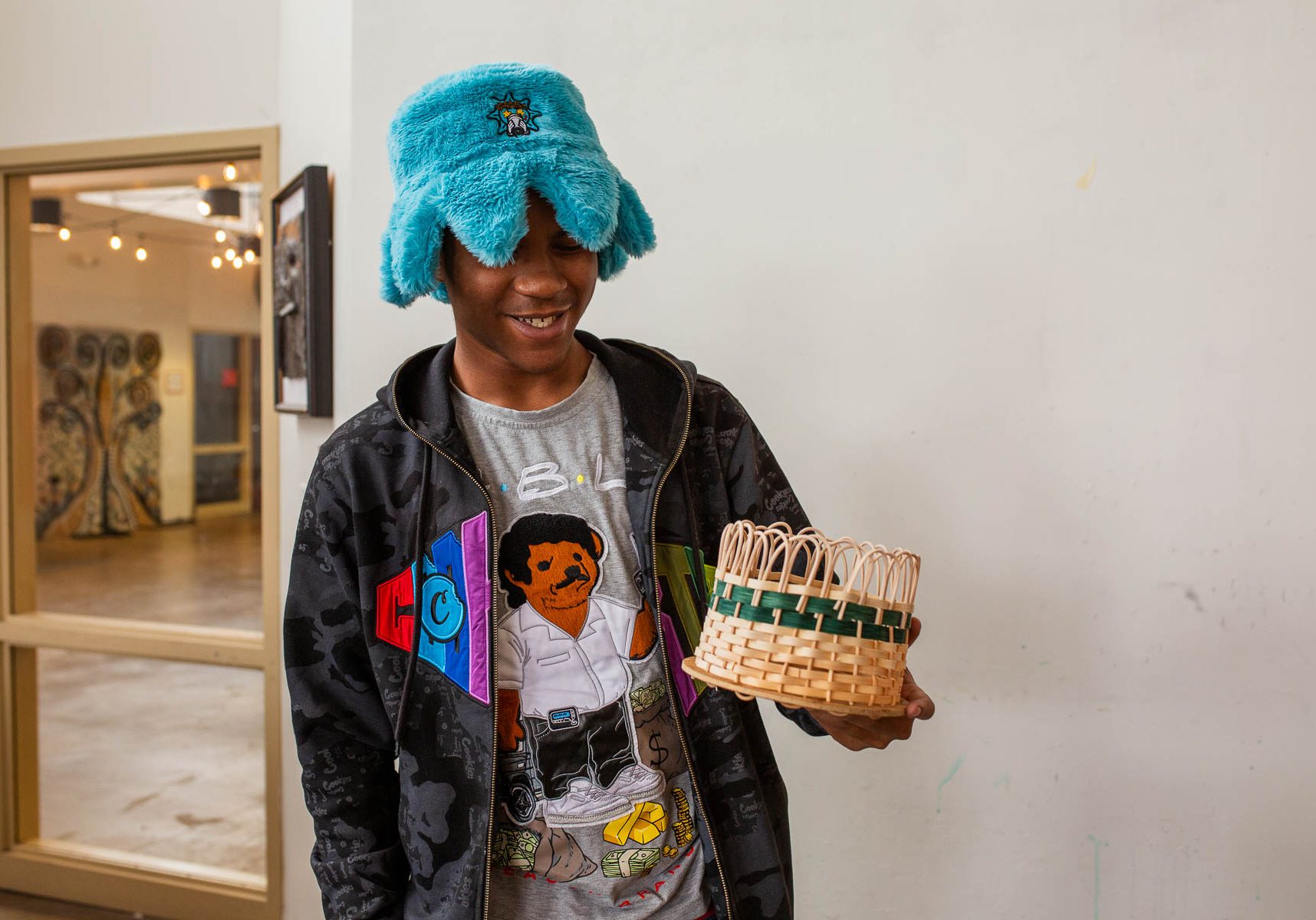 A smiling young person wearing a blue fuzzy hat holding a handmade woven basket.