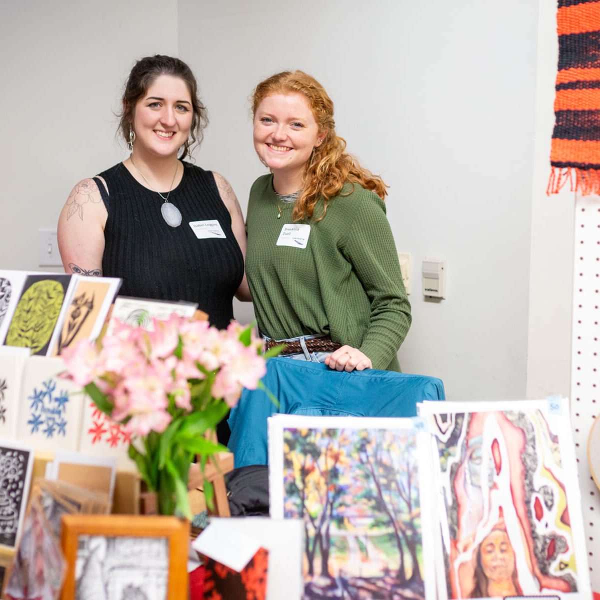 Two smiling women standing behind a craft display table.