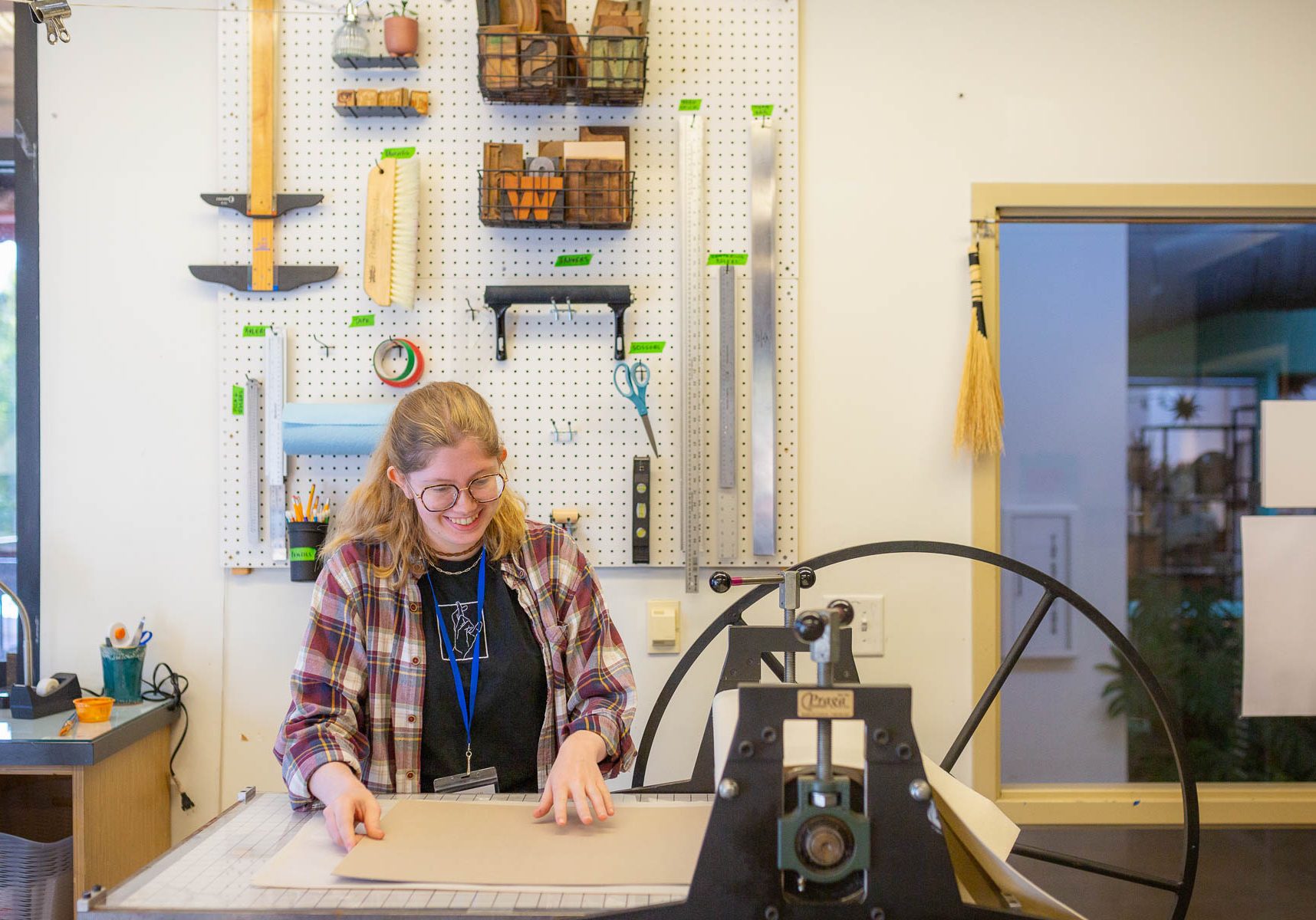 A smiling woman wearing glasses working at a table in a workshop or studio.