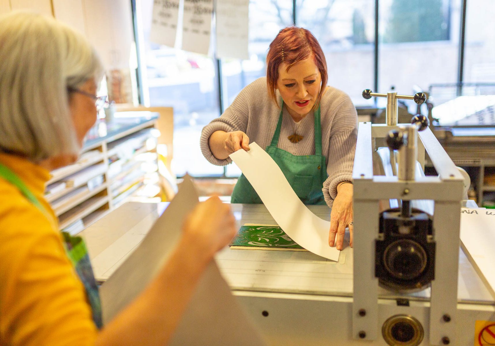 Two women smiling while holding up yellow and green printed art prints inside a bustling studio event.