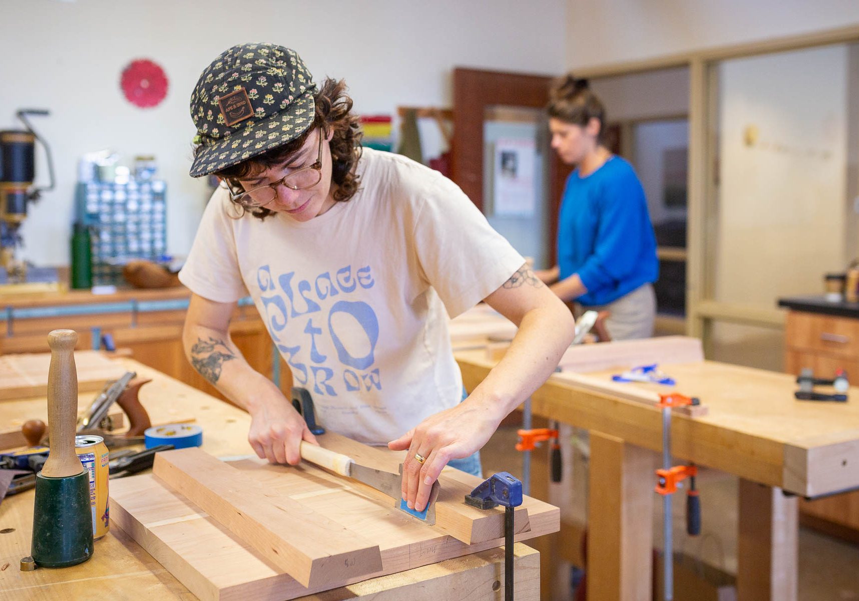 A person wearing a cap and graphic tee using a hand plane on a piece of wood at a workbench.