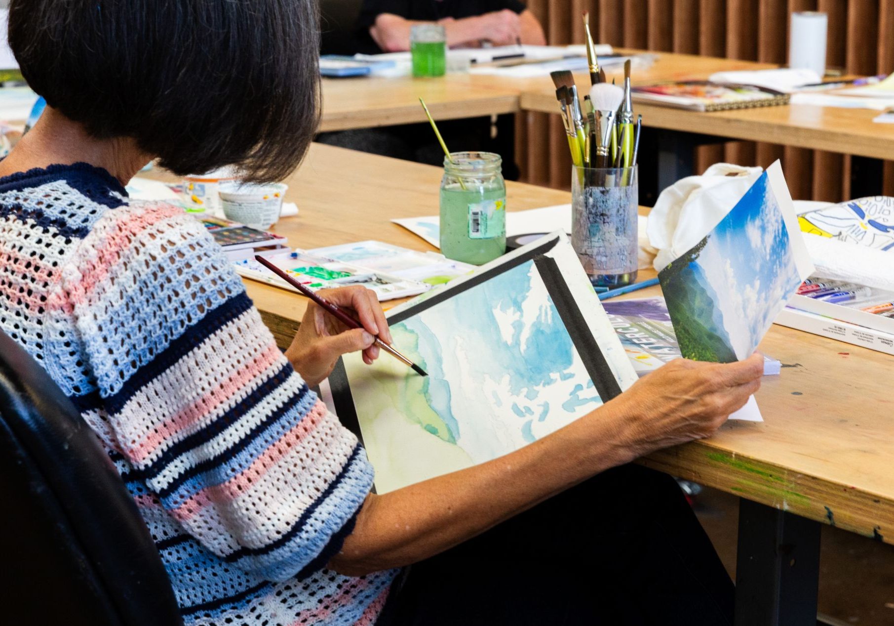 A woman painting a landscape in a sketchbook at an art studio table.