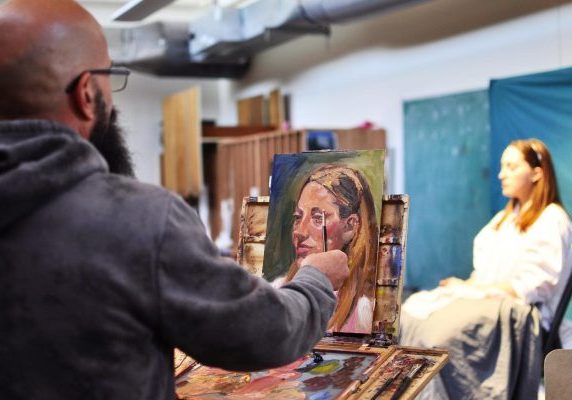 An artist painting a portrait of a model sitting in an art studio.