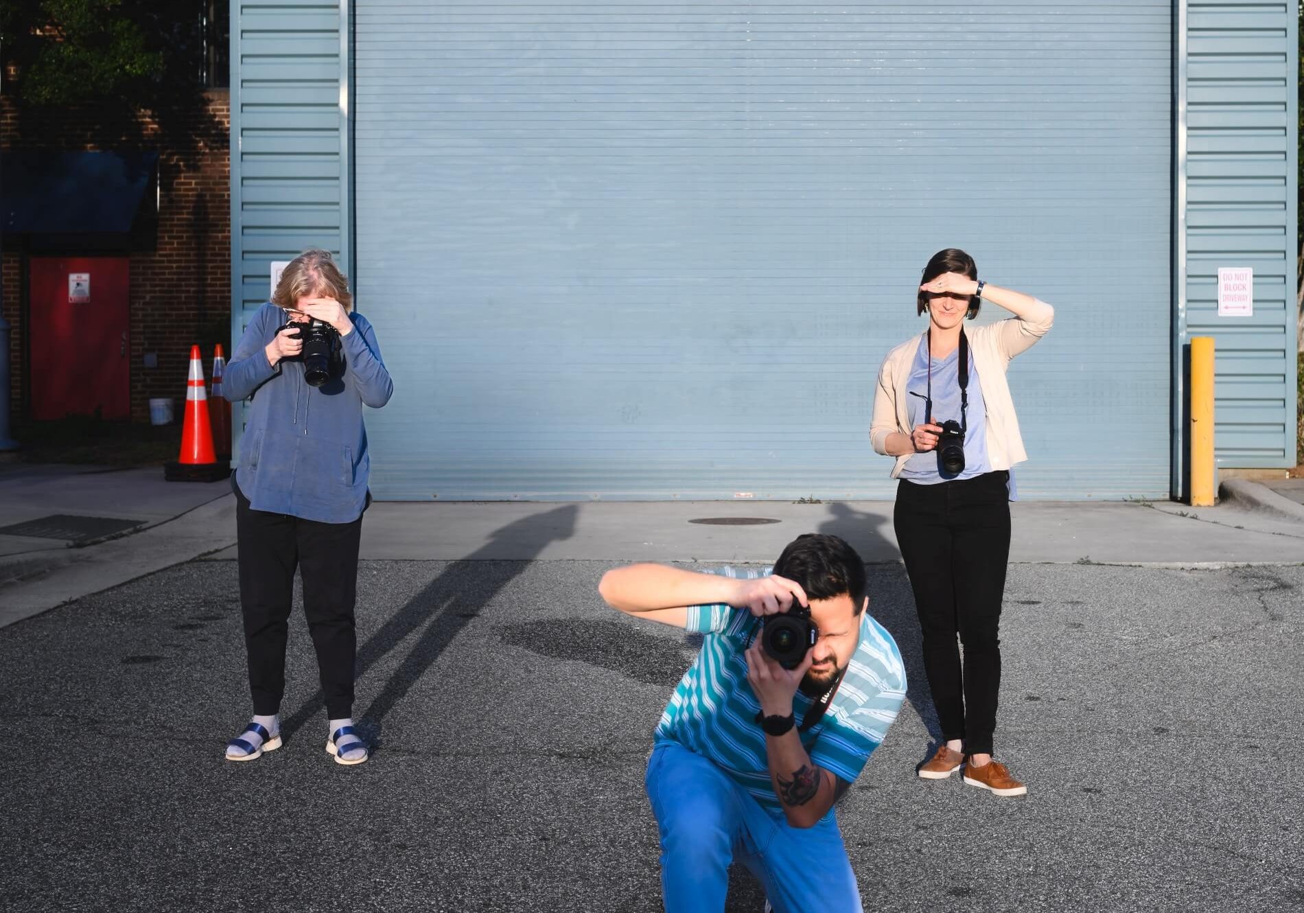Three photographers posing with cameras outdoors in front of a metal garage door.