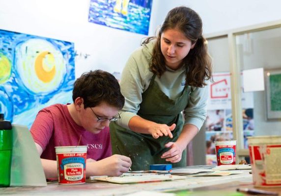 An instructor guiding a young student at an art studio table.
