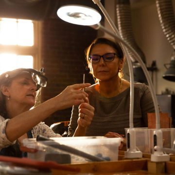 A woman using a desk lamp and tools at a workshop bench.