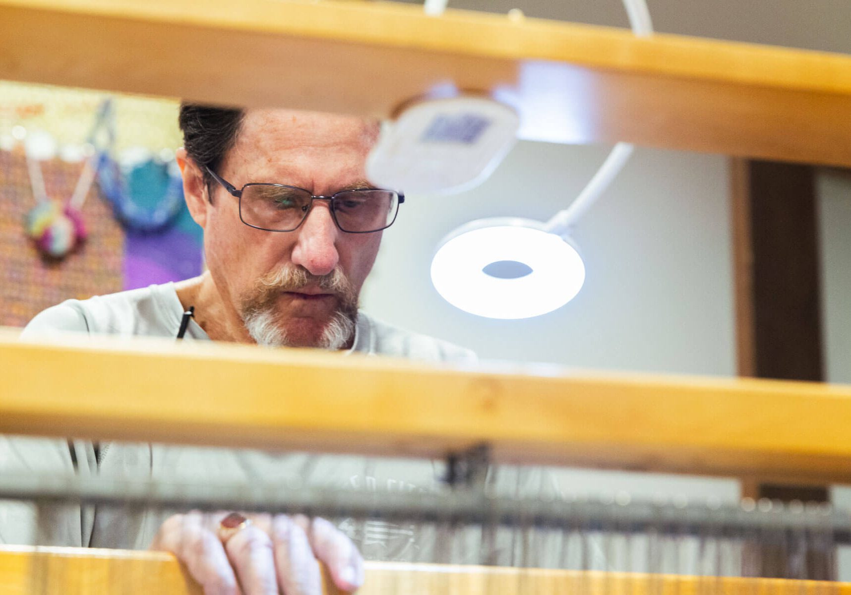 A man wearing glasses concentrates intently while weaving thread on a wooden floor.