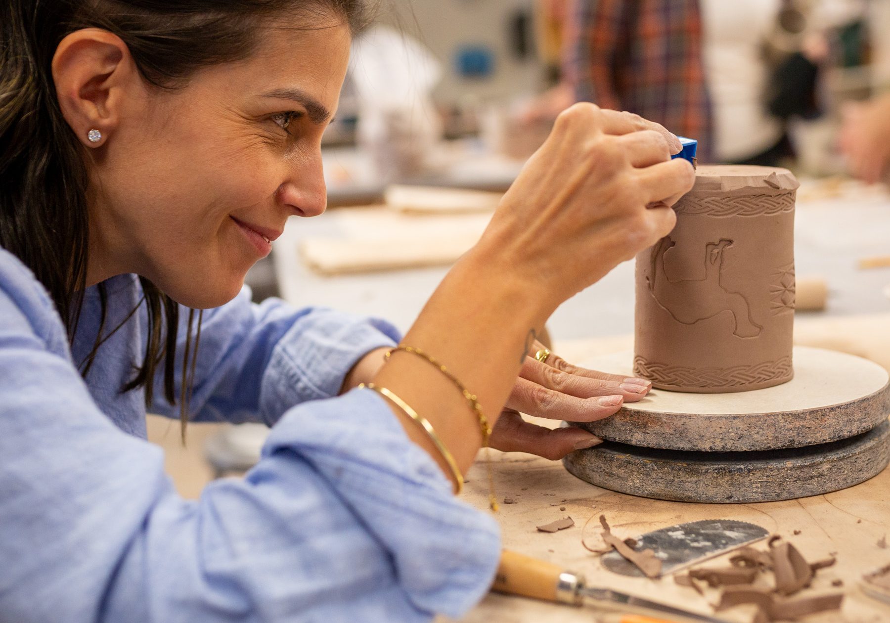 A woman carving details into a clay cylinder on a pottery wheel.