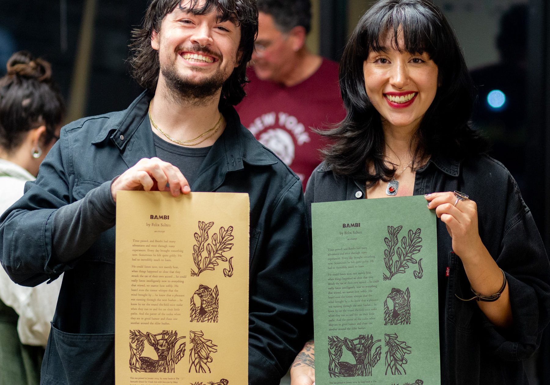 Two smiling artists displaying printed art posters in a studio.