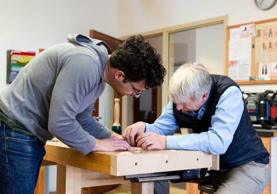 Two men collaborating on a woodworking project at a workbench in a shop.
