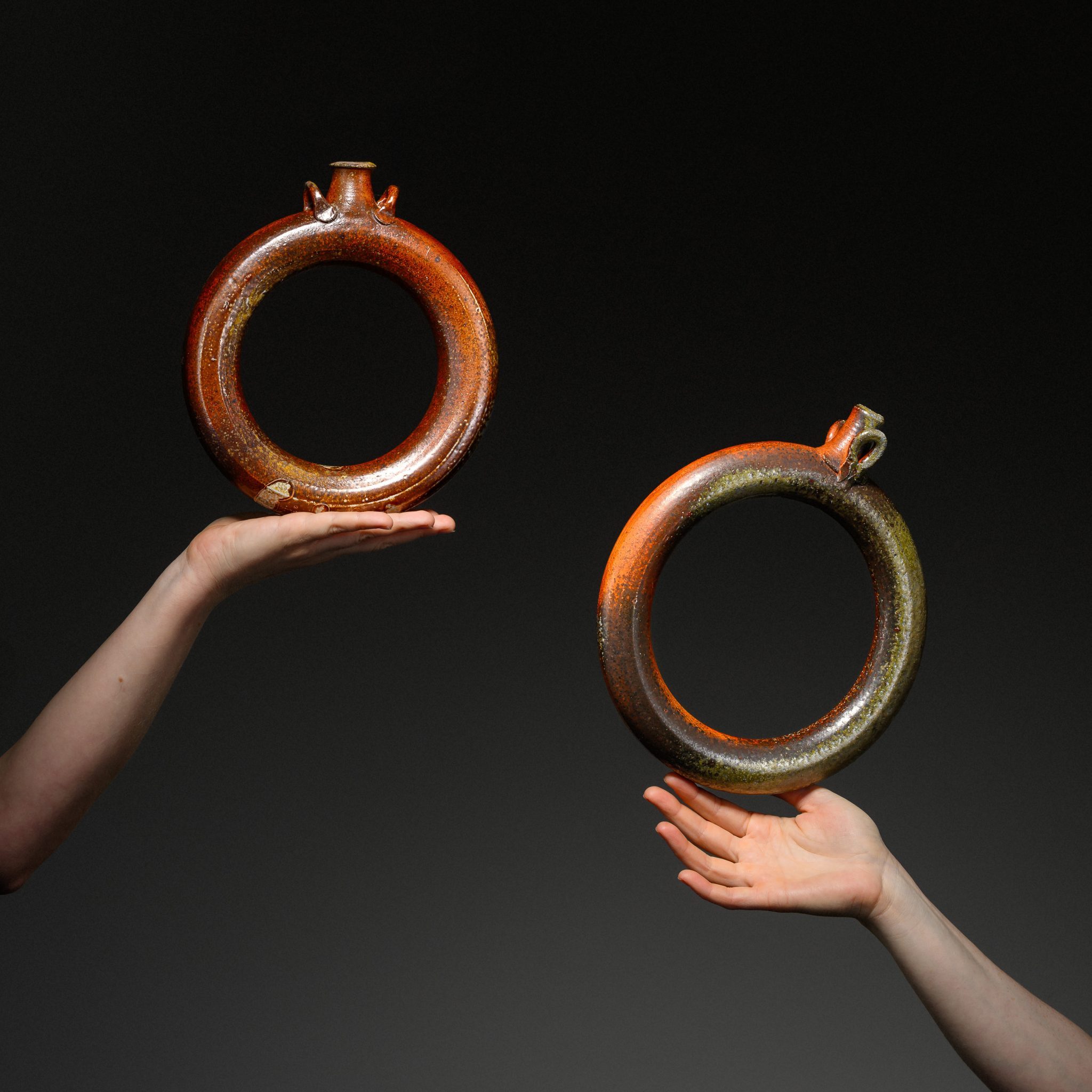 Two hands holding circular ceramic ring flasks against a dark background.