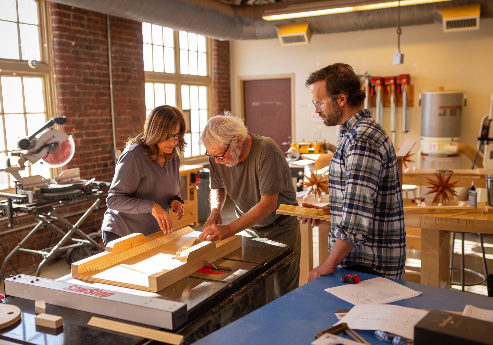 Three people working together at a table saw in a woodworking studio.