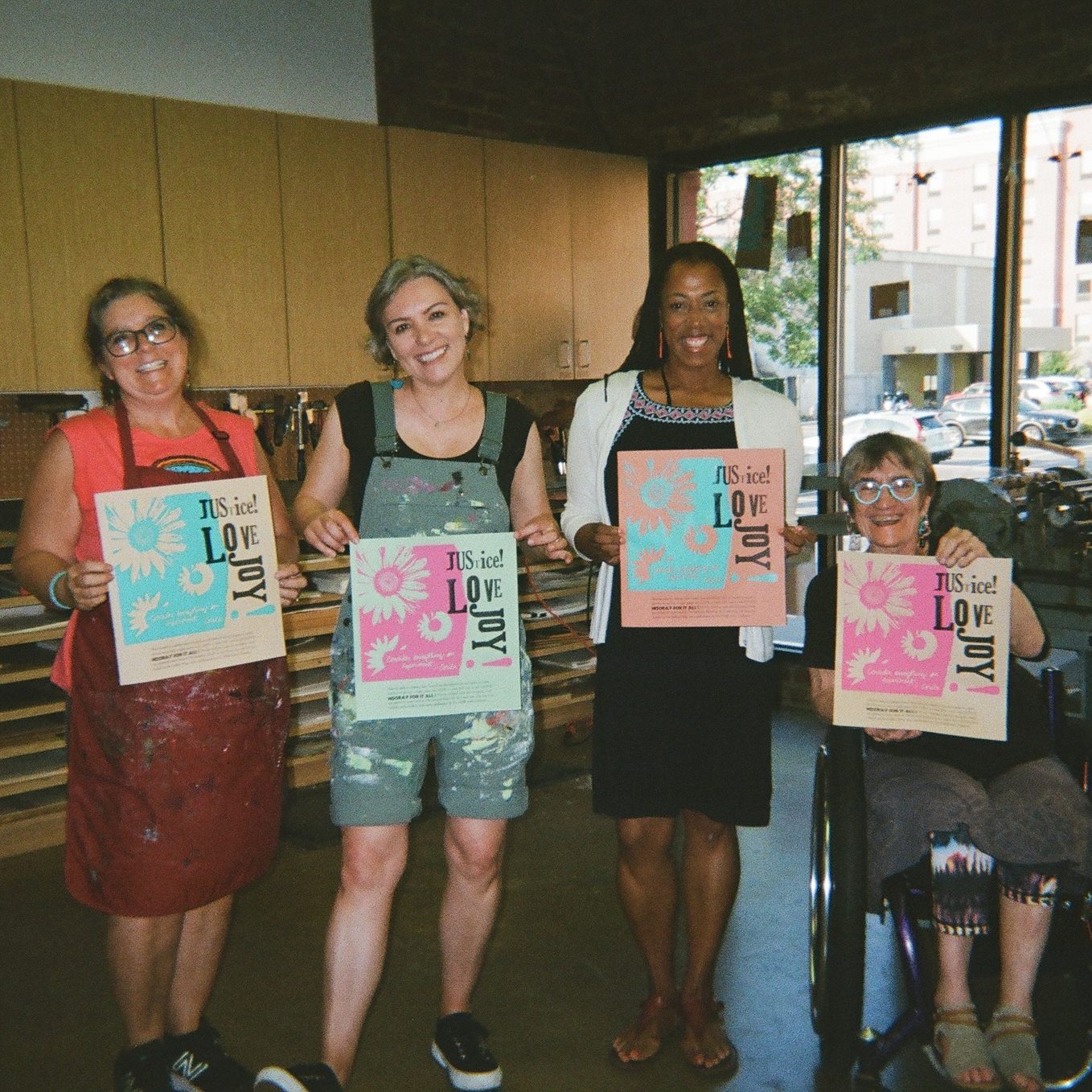 Four women holding up colorful art prints inside a bright printmaking studio.