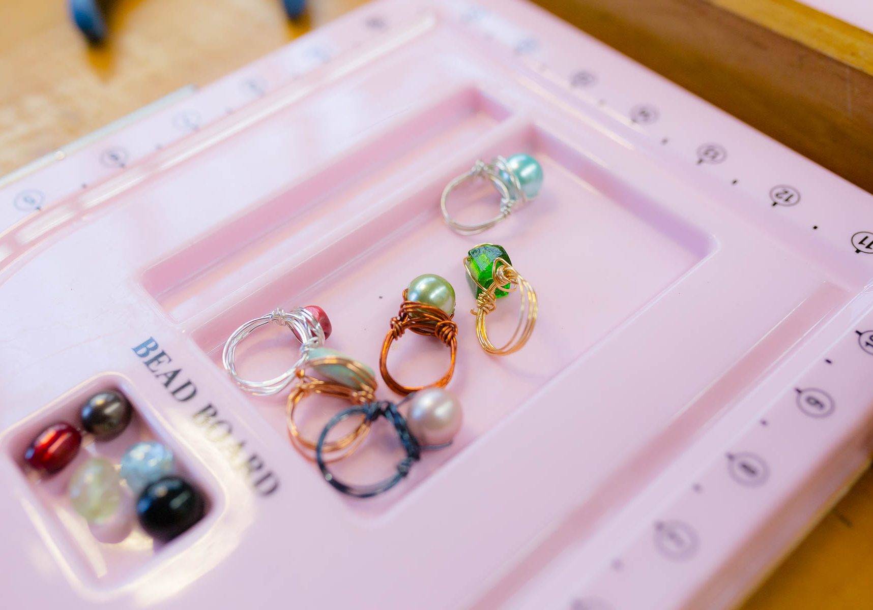 A collection of colorful wire-wrapped gemstone rings and loose beads displayed on a light pink bead design board.